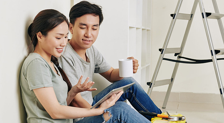 Man and woman sitting on floor in a white room with a ladder in the background. They talk while looking at an ipad in the woman's hand.