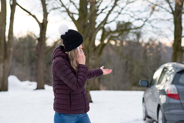 Woman standing in the snow talking on the phone, gesturing to parked car behind her
