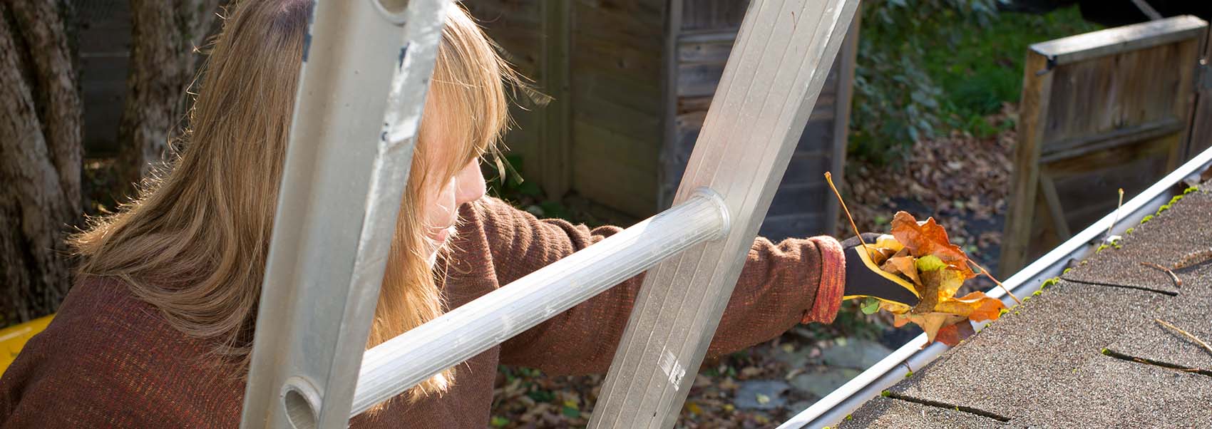 woman on a ladder cleans leaves and twigs from rain gutters or eavestrough