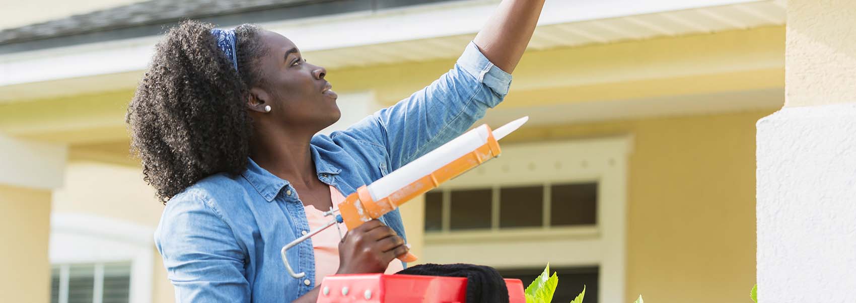 woman standing on a ladder outside her house, using a caulk gun to repair a leak