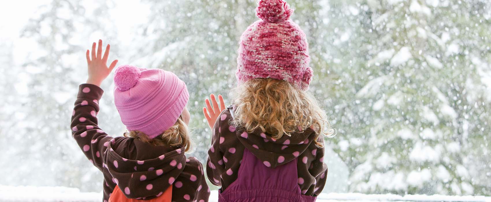 Two young girls in pajamas and knit hats looking out window at snow falling