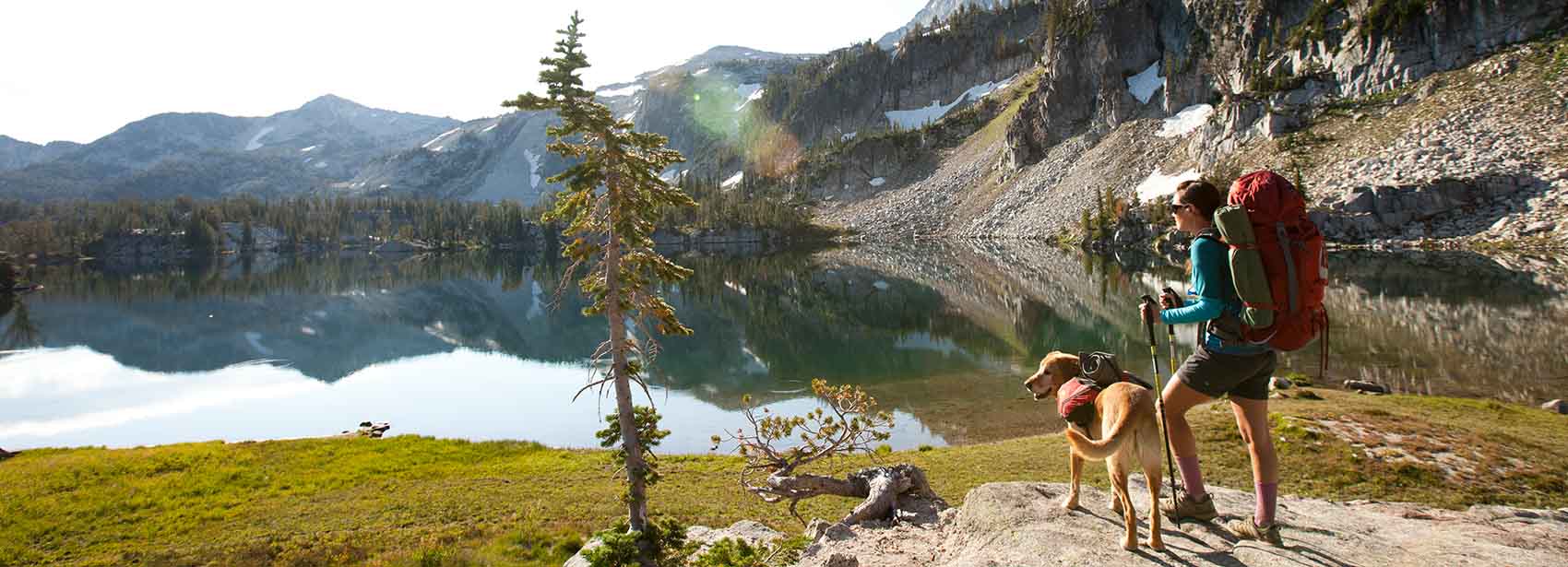 A woman and her dog stand on a large rock near a mountain lake at sunrise