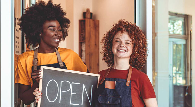 Two people smile outside of a small business, wearing aprons and holding an open sign.