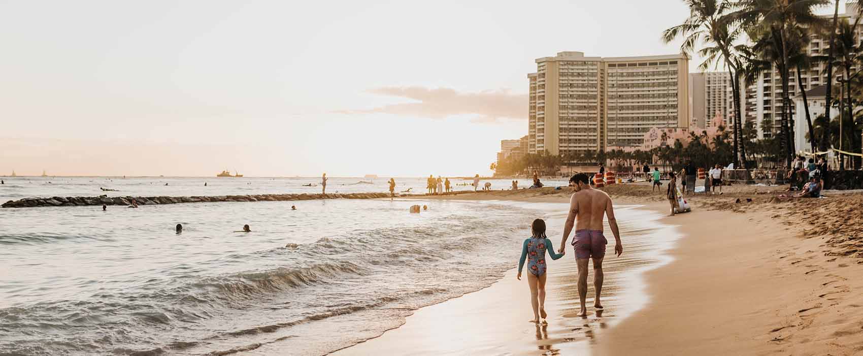 Father and daughter hold hands while they walk along beach at sunset