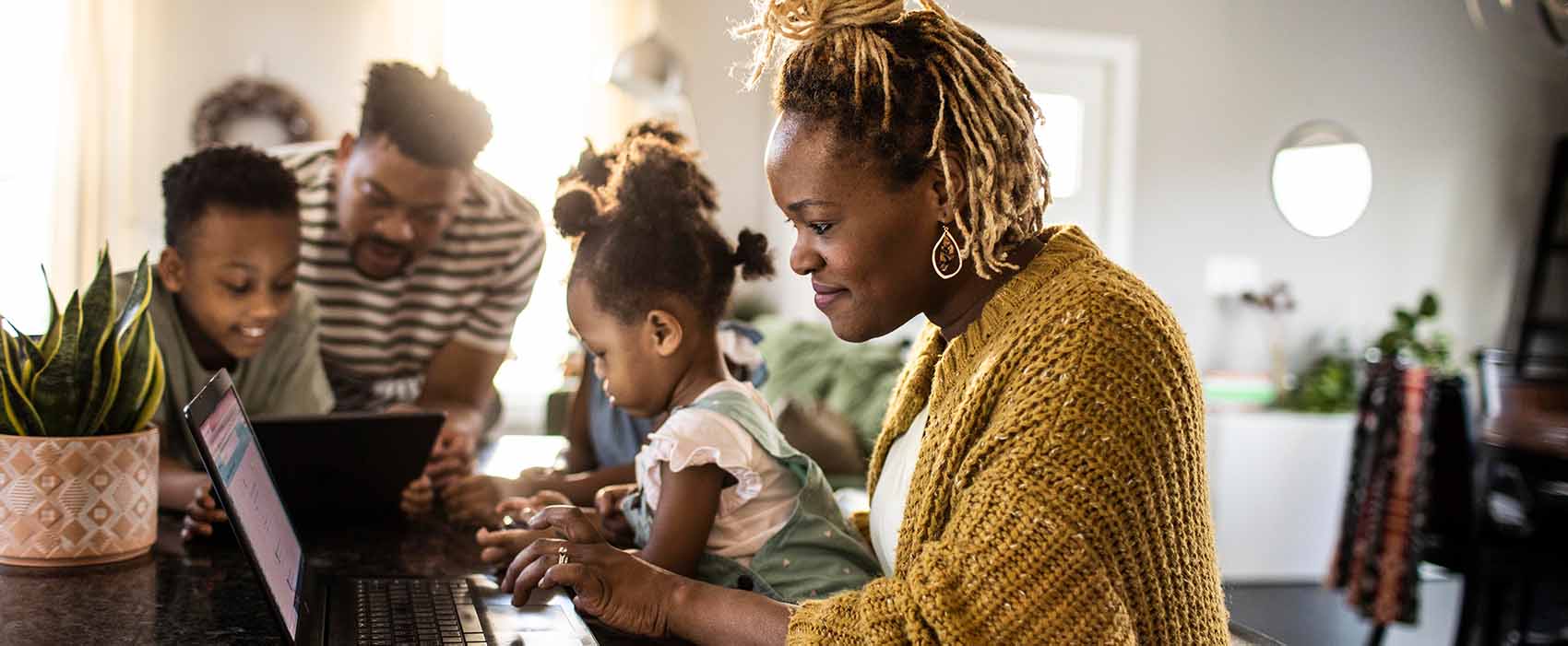 Mother working from home while holding toddler, family in background