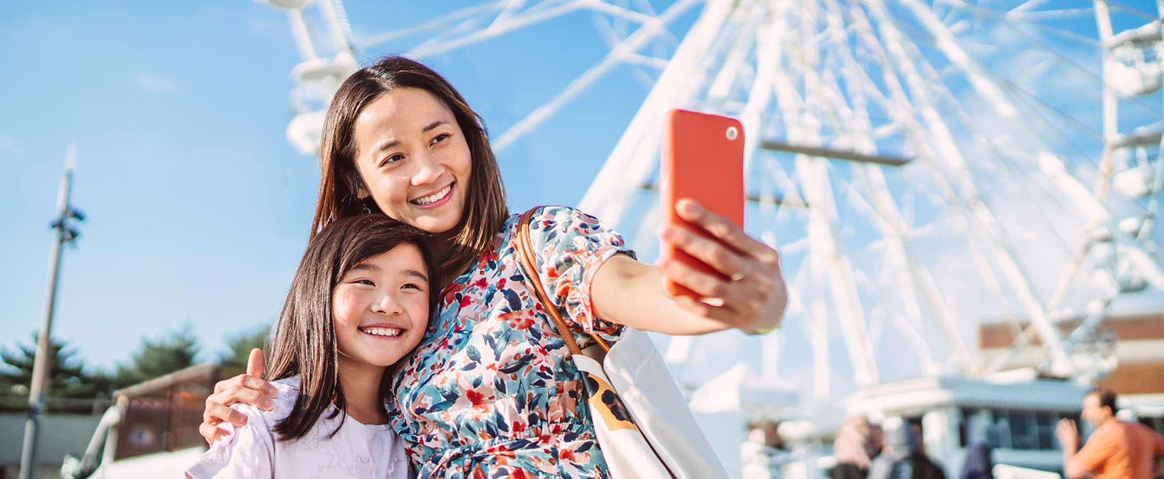 Mom and daughter take selfie with phone in bright orange case in front of a white ferris wheel on a sunny day