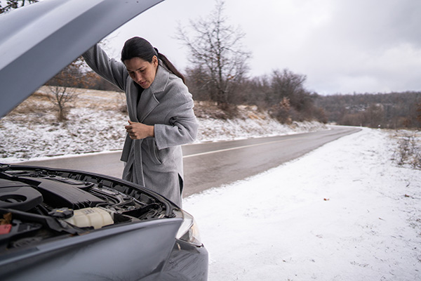 Woman looking under the hood of a car on the side of the road on a snowy day