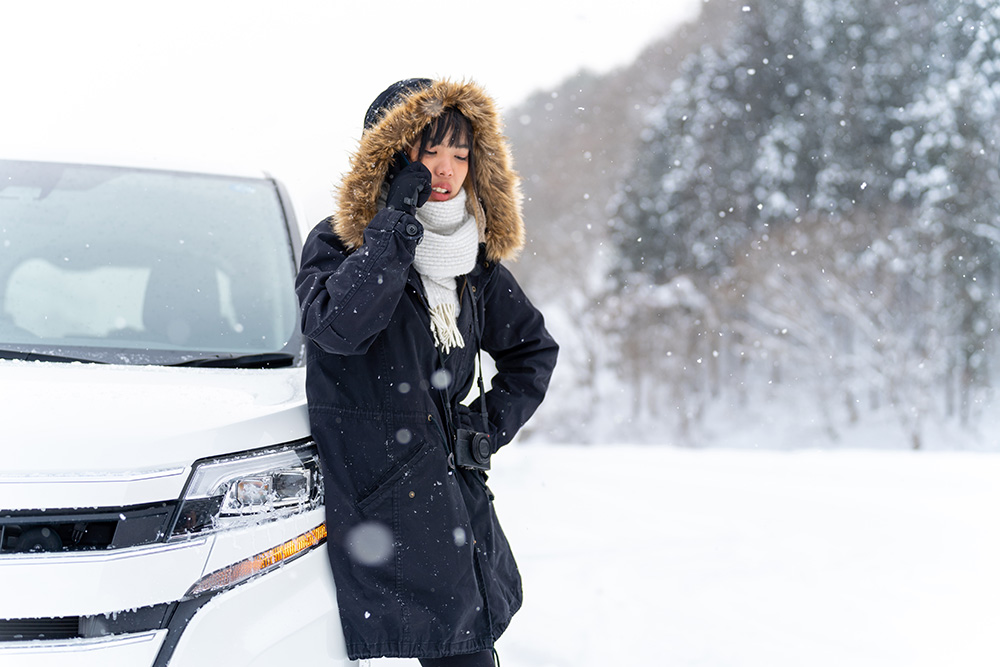 Woman leans against hood of white car in the snow, wearing a parka and talking on the phone.