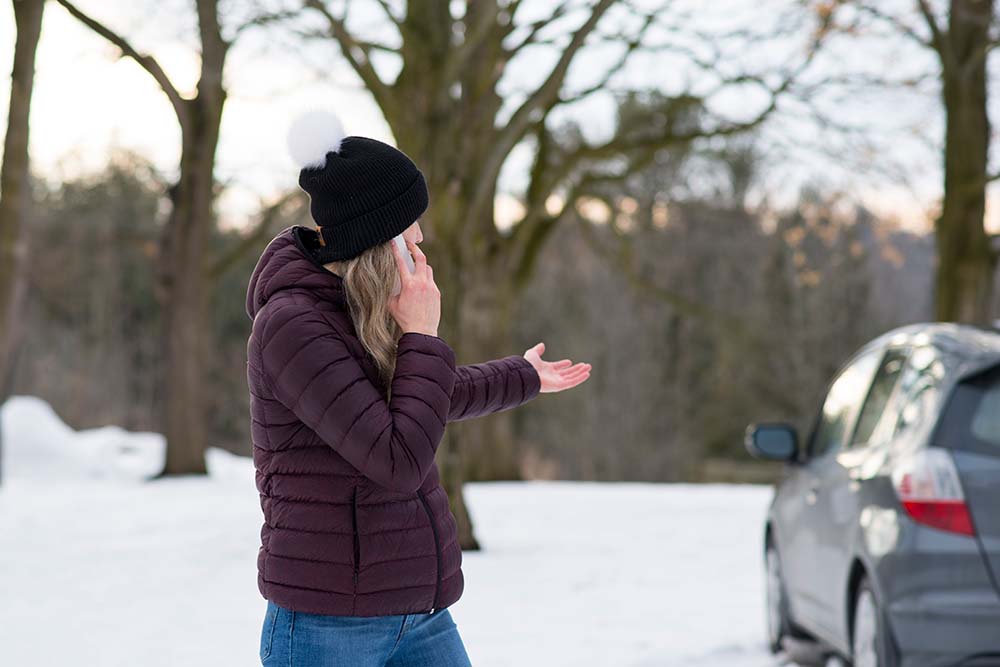 Woman standing in the snow talking on the phone, gesturing to parked car behind her