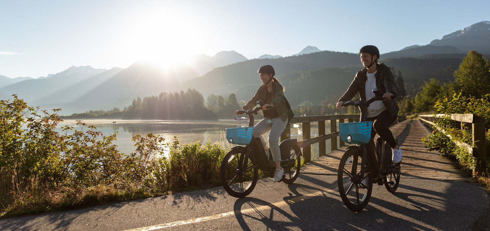 Two friends cycling over a wooden bridge with mountains in the background