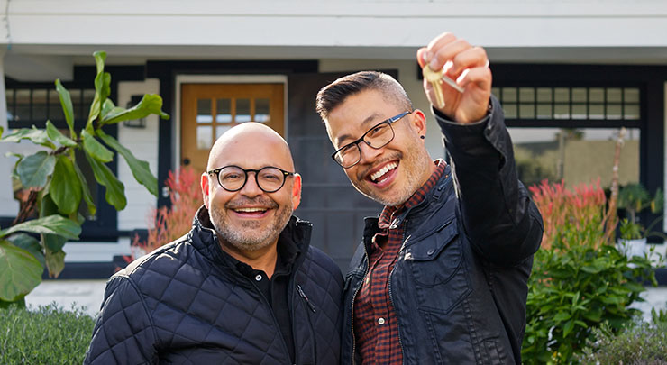Two men standing in front of a white home with black trim, smiling and holding keys up to the camera. 