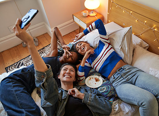 College friends on a bed, smiling for a selfie, with an empty plate placed next to them